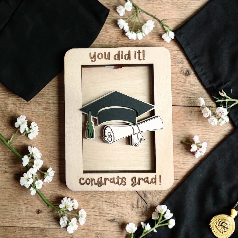 Graduation-themed wood gift card holder with 'you did it!' and 'congrats grad!' text, surrounded by flowers on a wooden surface.