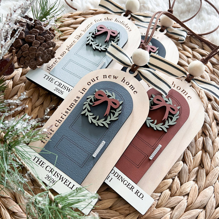 Three personalized wooden house-shaped gift tags with faux door designs and ribbon bows, arranged on a woven straw mat with pine branches and a pinecone.