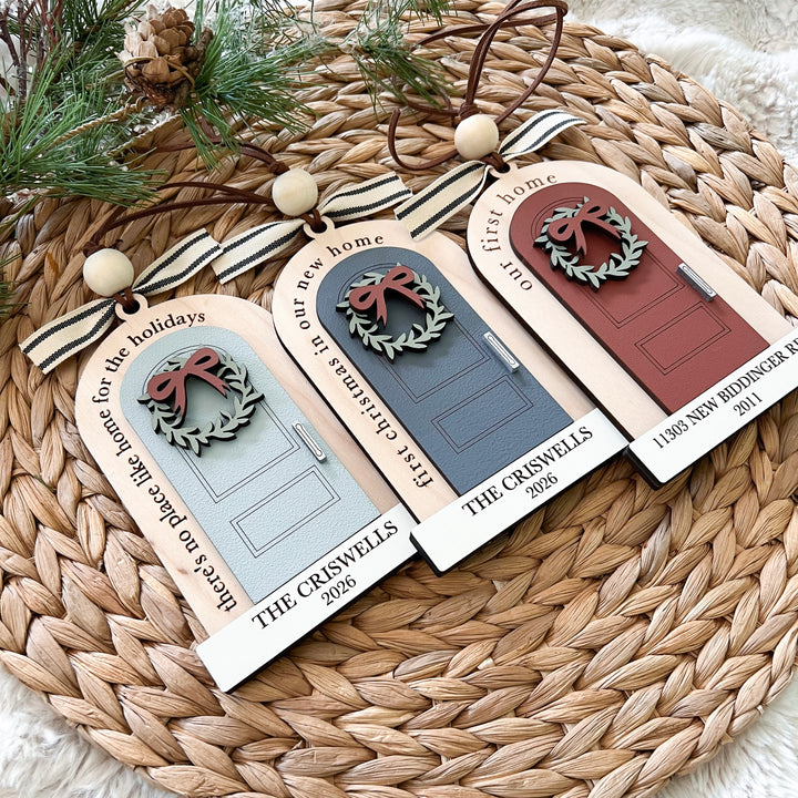 Three personalized Christmas door ornaments displayed on a woven straw mat with pine branches. Each ornament is shaped like a house door with a ribbon handle, bead accents, and printed text including 'our first home', 'our new home', and 'THE CRISWELLS 2026'.