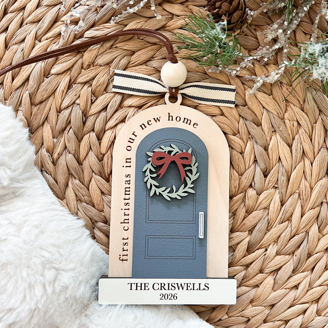 Personalized Christmas ornament shaped like a small house door with the text 'first christmas in our new home' and 'THE CRISWELLS 2026', hanging from a brown cord with a striped ribbon on a woven straw mat.