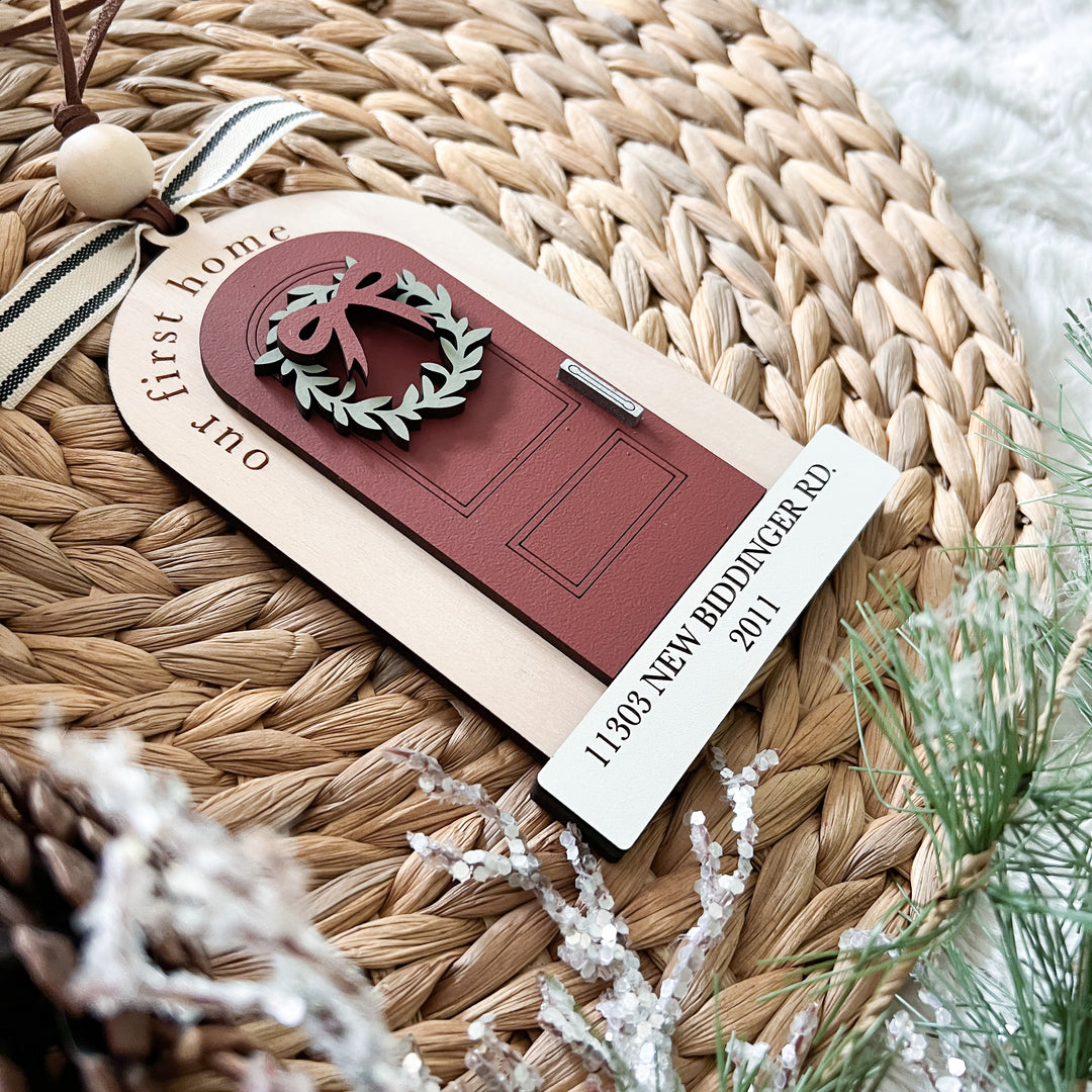 A personalized Christmas ornament shaped like a red front door with a green wreath and bow, attached to a beige card that reads 'our first home' and '11388 NEW BIDDINGER RD 2011.' The ornament is styled on a woven straw mat with ribbon, beads, and greenery around it.
