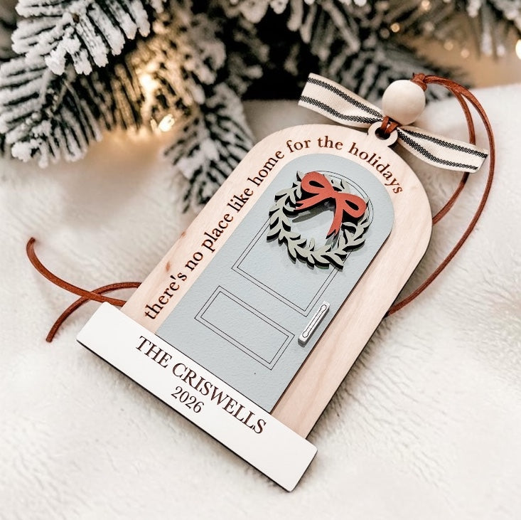 Personalized Christmas ornament shaped like a door, hanging with a ribbon and cord. The ornament has a gray door graphic with a red bow and wreath, the phrase 'there's no place like home for the holidays,' and a white hanging strip that reads 'THE CRISWELLS 2026.