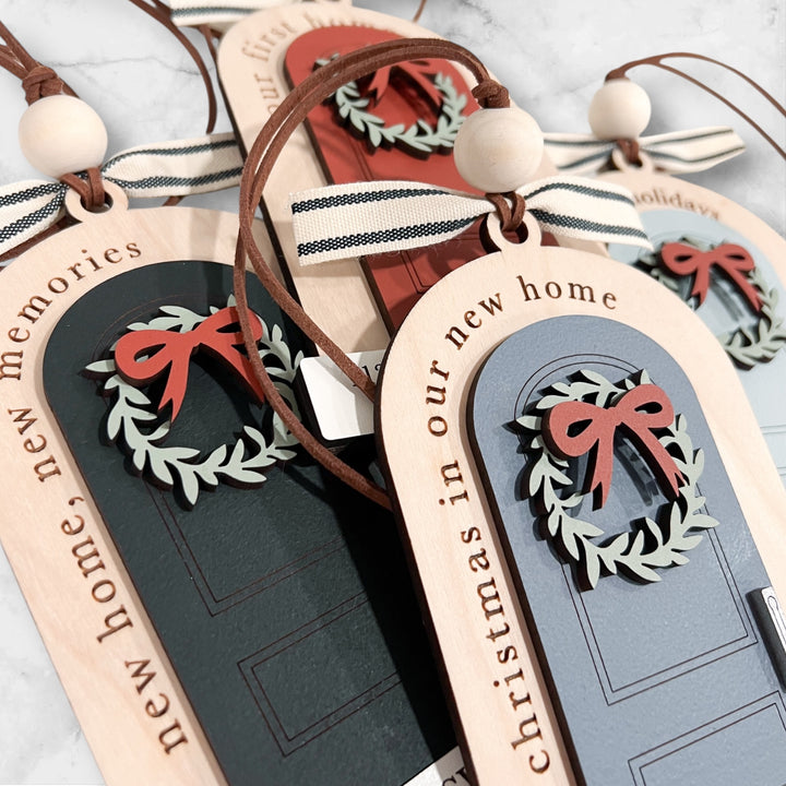 Close-up of several wooden Christmas door ornaments with ribbon hangers, laser-engraved door designs, and red bow details on a light marble surface.