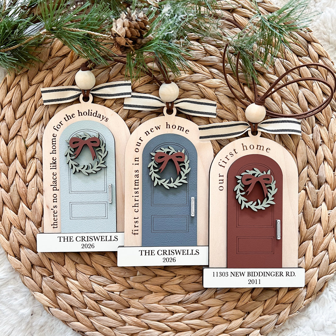 Three personalized wooden Christmas door ornaments displayed on a woven round mat with pine branches and a pinecone. Each ornament features a different door color: light blue, muted blue-gray, and reddish-brown, with a green wreath and brown bow.